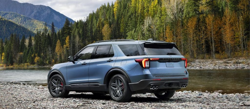 A blue Ford Explorer ST parked by a river surrounded by trees in a mountainous landscape.